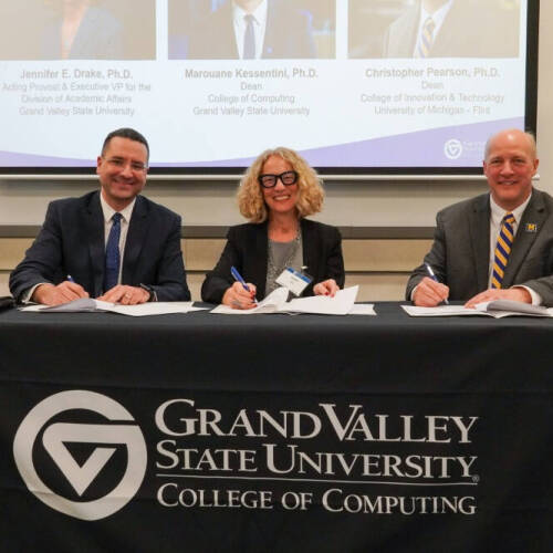Dean Kessentini, on the left, alongside other professionals sit along a table covered with a black GVSU College of Computing cloth. They hold pens as they sign an MOU while smiling to the camera